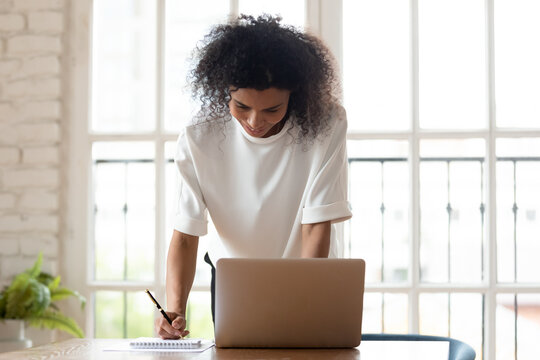Happy confident African American woman worker busy making notes browsing Internet using computer in office, biracial millennial female employee work on laptop write in notebook preparing report