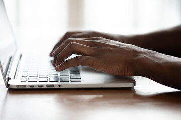 Close up of African American male employee busy typing texting on keyboard on computer, focused biracial man browsing Internet working on modern laptop on wooden desk indoors