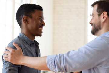 Smiling Caucasian businessman handshake tap shoulder of excited African American male employee...
