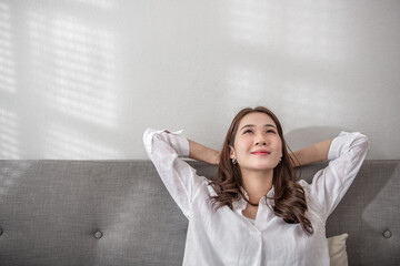 Asian young girl relaxing in living white room. Happy woman put hands behind head sitting leaned on couch at home with sunrise in the morning. Stress free and relaxation concept. Copy, space.