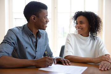 Excited young couple sit at desk put signature on contract feel euphoric buying first house together, smiling African American husband and wife look at each other sign agreement at realtor office