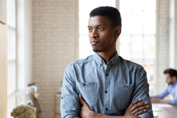 Pensive African American young male employee stand in office look in window distance thinking, thoughtful biracial millennial man worker pondering planning, business vision concept