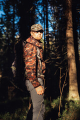 Fototapeta na wymiar Hiker hiking in autumn forest. Male hiker in camouflage jacket with backpack looking to the side walking in forest. Caucasian handsome male outdoors in nature. Concept of forest wear, navigation.