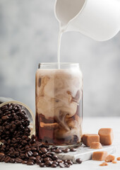 Pouring milk into glass with iced black coffee with glass jar of coffee beans and salted caramel on light background.