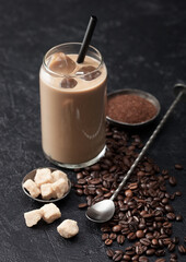 Iced coffe in glass with straw with beans and cane sugar with ground coffee and long spoon on black.