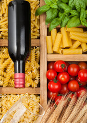 Wooden box with various classic italian pasta and basil with tomatoes and bottle of red wine.