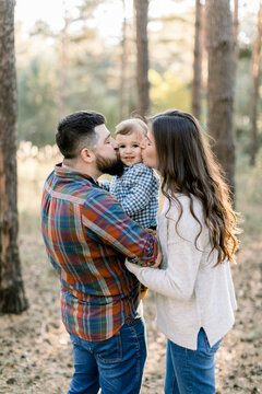 Lovely Caucasian family, handsome bearded father, pretty brunette woman mother, and cute little baby son, having fun while walking in an autumn pine forest. Parents kissing their baby outdoors
