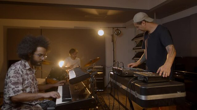 Music production studio. Band rehearsal. Hispanic man playing keyboards, caucasian man playing drums, young man doing beats. Talented musicians record their ideas. 