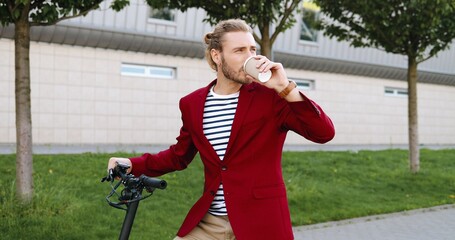 Caucasian young handsome man in red jacket standing on street at bike or electric scooter and drinking coffee. Stylish guy sipping drink to-go outdoors. Bicycle. Bicyclist stop to rest.