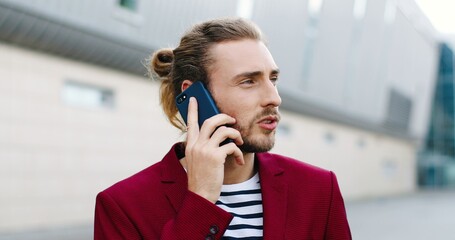 Close up of young handsome Caucasian man in red jacket talking on mobile phone outdoors. Serious good-looking stylish male standing at street and speaking on cellphone. Telephone conversation.