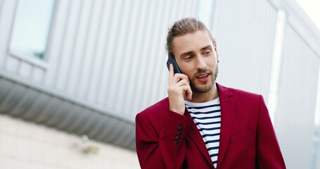 Young handsome happy Caucasian man in red jacket talking on mobile phone outdoor. Cheerful...