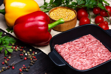 Minced pork in a black plate on a wooden table along with spices and bell pepper, cherry tomatoes, parsley