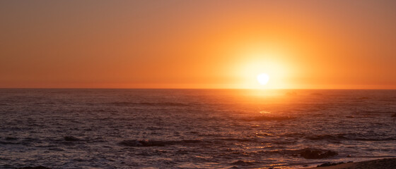 Naklejka premium Bright vivid sun on horizon over ocean at sunset, with reflections on water. Wide aspect ration with copy space on left.
