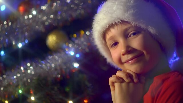 4K video of a smiling boy  is praying near Christmas tree about christmas gifts. Boy in a Santa hat is dreaming Christmas gifts on  Christmas.  Happy 8 year old child is preparing for the New Year.