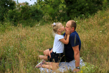Naklejka na ściany i meble Three-year-old blonde daughter kisses dad on the nose, gently hugging his cheeks. Dad and daughter are sitting in the grass in nature. Father's love for his daughter. Family vacation in nature.