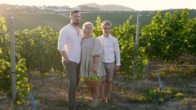 family of three in white clothes stands in a vineyard at sunset