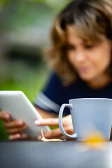 Beautiful, young woman using electronic book reader on a table outside with a cup of coffee in focus