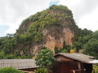 A large limestone mountain with some trees. There are wooden houses nearby at Ban Jabo, Mae Hong Son, Thailand.