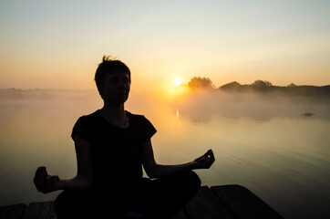 Silhouette of a meditating girl against the background of sunrise with copy space. Woman in kubera mudra meditation. Reflection of the sun in the water surface.