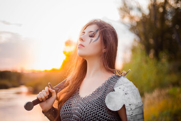 Portrait of a medieval woman warrior in chain mail with a sword and shield in her hands against the backdrop of the river and sunset. Combat makeup, clear skin, retouching.
