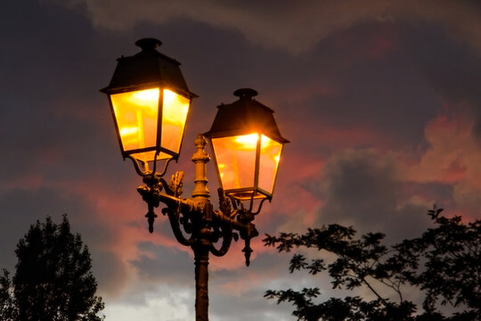 Vintage street lamp, slightly glowing with orange light in the rosy dusk, with trees silhouettes in the background