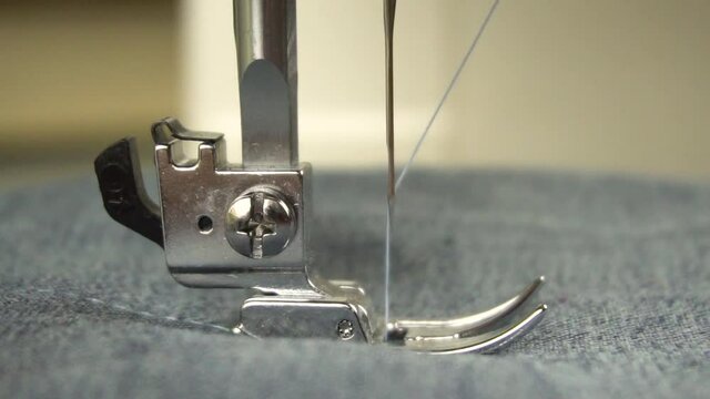 Slow motion of the foot and needle while sewing on the sewing machine.The silver presser foot of the sewing machine presses the fabric and sews it with a needle.