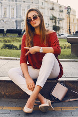 Pretty stylish  woman in trendy sunglasses and red sweater sitting and   flirting . Outdoor street style portrait. Cheerful woman wearing  heels and white jeans.