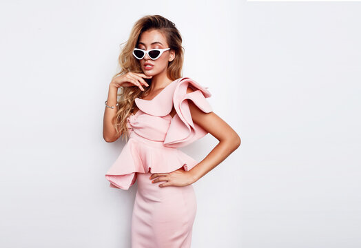 Fashion studio image of gorgeous elegant woman in pink dress and sunglasses posing over white wall. Blonde wavy hairstyle. Sext full lips.
