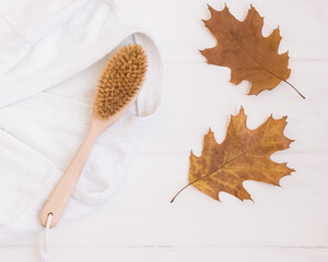 Wooden body brush, bathrobe and autumn leaves on a white wooden background. Selective frocus on the...