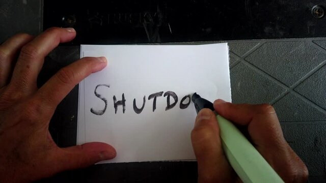 Adult Caucasian female hand writes with black magic marker and scribes in block capital letters word shutdown on white piece of square paper, covid-19 quarantine and pandemic, directly above static