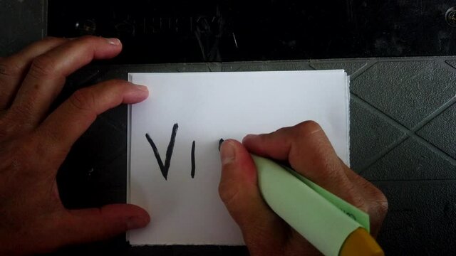 Adult Caucasian female hand holding black magic marker scribes in block letters the word virus on white piece of square paper, covid-19 pandemic and quarantine, directly above static