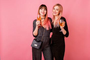 Stylish celebrating women in party dress drinking shampagne and have great time together. Pink background. Happy emotions. Fashionable outfit.