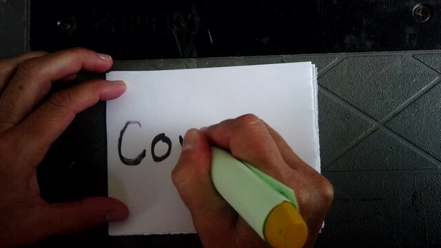 Adult Caucasian female hand holding magic marker scribes in black letters the word covid-19 on white piece of square paper, directly above static