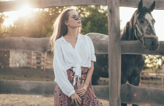 
a woman of model appearance with long hair is dressed in a shirt with a long skirt and holds a guitar in her hands stands near the fence against the background of an equestrian farm near the horses