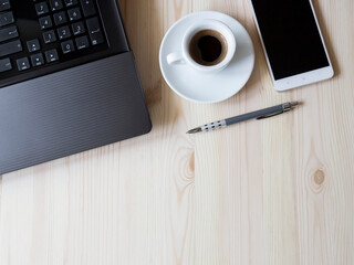 Top-down shot of part of a black laptop, black coffee in a white cup, a white smartphone with a black screen and a pen on a table. The table is made of light wood. Half of the tablespace is empty