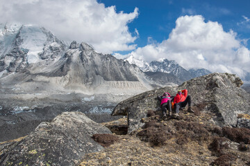 A couple of trekkers are lying resting on a big stone under the sun in the Himalayas. A glacier and snow-capped mountains in the background.