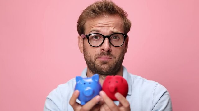 clueless young man wearing glasses, holding piggy banks and examining, pondering and shrugging on pink background