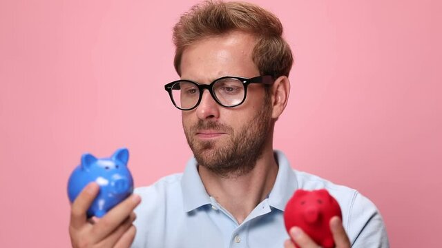 unsure young casual man in blue polo shirt holding piggy banks, analysing and shrugging on pink background