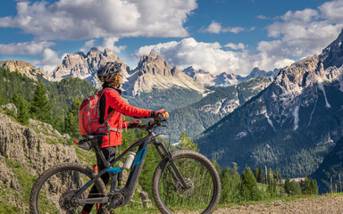 nice and active senior woman riding her electric mountain bike on the high plateau of Pratto Piazzo in the three peaks Dolomites , rocky silhouette of Mount Cristallo in background, South Tirol, Italy