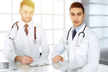 Fototapeta premium Arab doctor man sitting at the glass desk with caucasian colleague in medical office or sunny clinic. Diverse doctors team, medicine concept