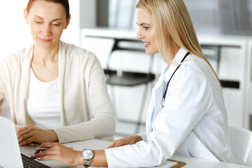 Obraz premium Woman-doctor at work in sunny hospital is happy to consult female patient. Blonde physician checks medical history record and exam results while using clipboard
