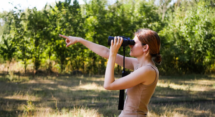 Obraz premium Woman with binoculars looking for animals in the park. Forest hiking banner. Bird watching. Tourist exploring wild nature. Points to the distance