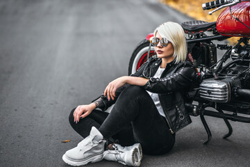 Obraz na płótnie Canvas Pretty blonde biker girl in sunglasses sitting near red motorcycle on the road