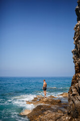 A young man stands on the rocks overlooking the open Mediterranean Sea. A guy on a warm summer sunny day looks at the sea breeze