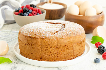 Homemade classic vanilla sponge cake or biscuit sprinkled with powdered sugar and fresh berries on a white plate on a light wooden background.