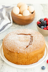 Homemade classic vanilla sponge cake or biscuit sprinkled with powdered sugar and fresh berries on a white plate on a light wooden background.