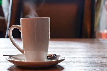 Tall white coffee mug on a saucer with a teaspoon on a wooden table in a cafe. Steam over a mug from a hot drink. The concept of a cozy breakfast. Space for text.