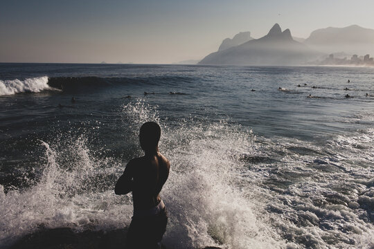 Man standing on the coast rocks behind splashing wave looking at a group of surfers in the sea waiting for the perfect wave in Rio de Janeiro, Brazil. Freedom lifestyle covid reopening concept.