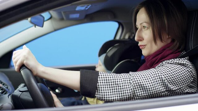 Brunette woman in casual clothes is driving car with greenscreen background. She is sitting in her car with her son on the bluesceen background isolated. her son is drinking baby food