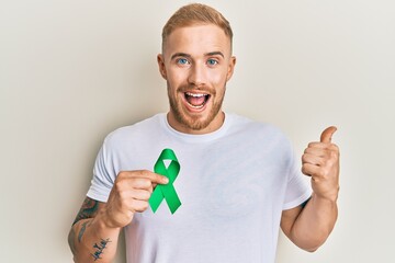 Young caucasian man holding support green ribbon pointing thumb up to the side smiling happy with...